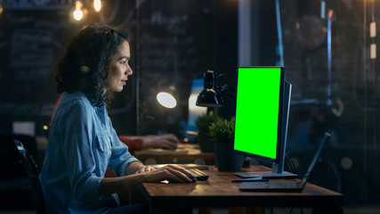 Wall Mural - Beautiful Female Office Employee at Her Desktop Works on a Mock-up Green Screen Personal Computer. Her Colleague Sits Beside. Creative Office Evening.