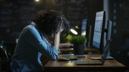 Tired, Overworked Female Mobile Phone App Designer Holds Her Head in Hands while Working on a Personal Computer. In the Background Creative Office.