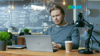 Canvas Print - Handsome Young Office Employee Thinks on a Problem Solution While Typing on a Laptop Computer. He's Working in the Creative Stylish Office.