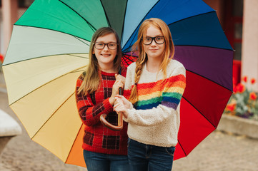 Group of two cute little girls playing outside under big colorful umbrella, wearing glasses and warm pullovers