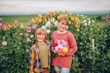 Outdoor portrait of two funny kids plying in autumn garden, holding dahlia flowers bouquet