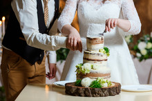 Groom In Suit And Bride In White Dress Cut Beautiful Multi Level Naked Wedding Cake, Decorated With Cream And Fresh Flowers, Greenery. Delicious Dessert At Wedding Banquet Standing On Wooden Plate
