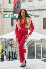 African American Woman Fashion in New York. Young woman with long hair dressing in red slim fashionable jacket, pants, black high heel pumps shoes, faux fur scarf on shoulder, standing on Wall Street