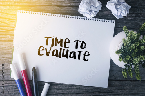 Fotomural Time to evaluate word with Notepad and green plant on wooden background