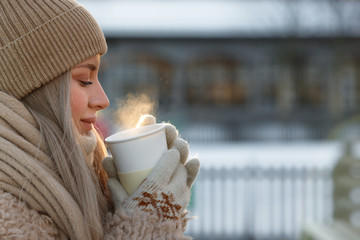 Young woman in beige fur coat, hat with pompon, scarf and white mittens holding steaming white cup of hot tea or coffee, outdoor in sunny winter day, close up/ Winter time concept/ Bask in the cold