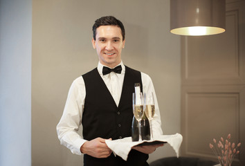 Canvas Print - Young waiter holding tray with glasses and bottle of champagne indoors