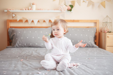 Portrait of cute adorable Caucasian blonde smiling baby girl in white onesie sitting on bed in bedroom and holding pink flower rose. Happy childhood lifestyle concept