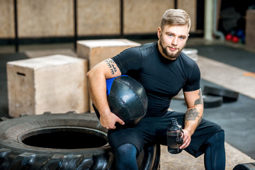 Portrait of a handsome athletic man in black sports wear sitting with ball on the tire in the crossfit gym