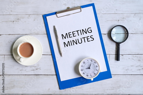 Fotografía Top view of clipboard with paper written 'MINUTES MEETING' with magnifying glass,pen,table clock and a cup of coffee on white wooden background