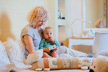Curly Blond Caucazian Grandmother With Her Grandson Sitting On Bedroom And Playing With Alarm Clock. In Cozy Home Interior. Real People Life And Different Generations Concept.