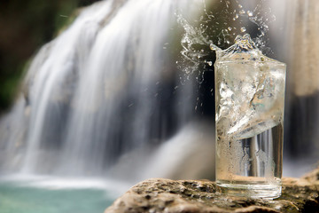 Dewy glass full of water with ice cube stands on a rock near a waterfall in the forest.