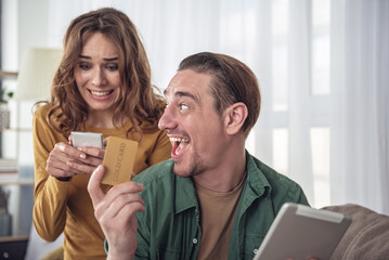 Excited man is showing cold credit card to his wife and laughing. Woman is typing on smartphone quickly while ordering things in internet. Online shopping concept