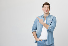 Studio Portrait Of Attractive European Man In Denim Glothes, Pointing At Upper Left Corner With Index Finger, Expressing Excitement, Happiness And Surprise, Over Gray Background.