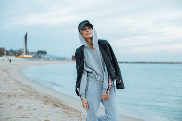 Happy and natural beauty young pretty and attractive woman or teenager wears grey hoodie and sweatpants during workout at beach seaside laughs naturally to camera, during fashion photoshoot influencer