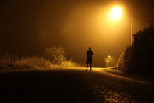 The Silhouette Of A Man In Shorts, Standing In The Middle Of The Road On A Misty Night. The Glare Of The Street Light Against The Fog Sets A Creepy Mood. Concept Of Thieves, Rapist And Killers Roaming