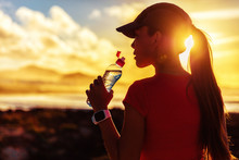 Fitness Woman Drinking Water From Sports Bottle On Afternoon Workout After Run Training Jogging Outdoors At Sunset. Girl Wearing Running Cap Silhouette Against Sun Flare.