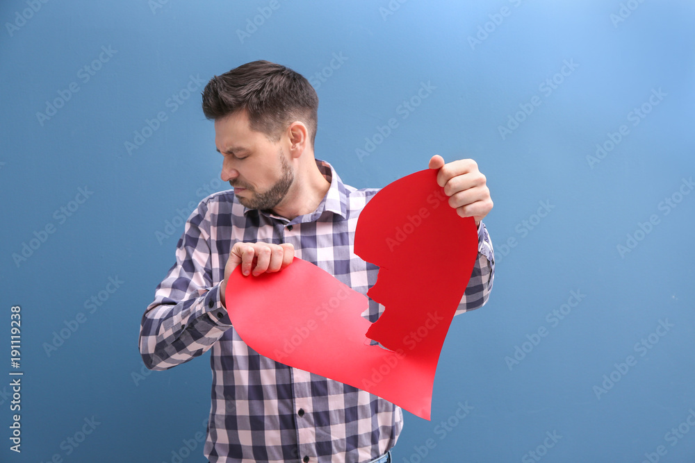 Young man tearing paper heart in half on color background. Relationship problems