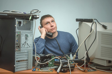 Wall Mural - Tired and bored computer technician engineer is sitting on his workplace and consulting a users by a phone. Computer repairman tired from his work. PC repair service center.