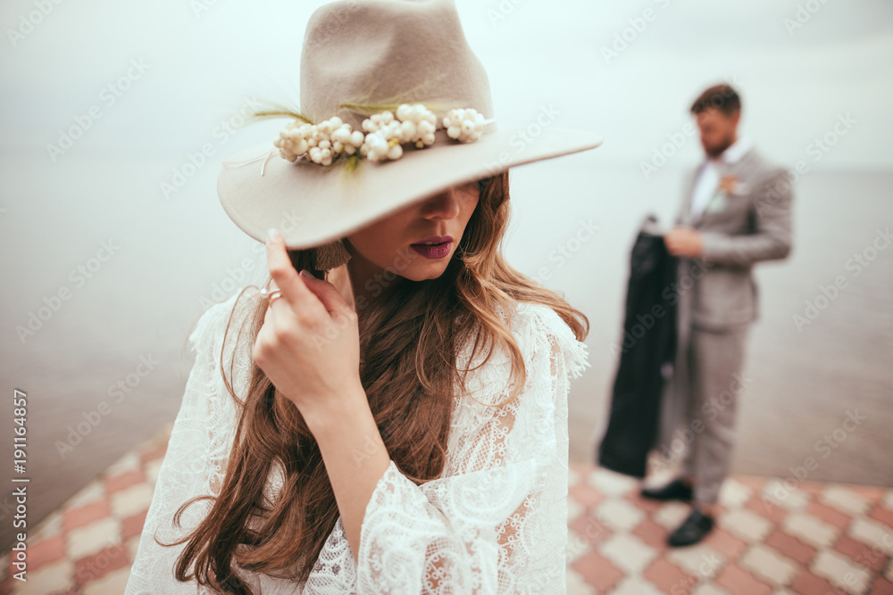 Obraz na płótnie beautiful bride in wedding dress and hat in boho style on pier at lake, groom standing behind w salonie