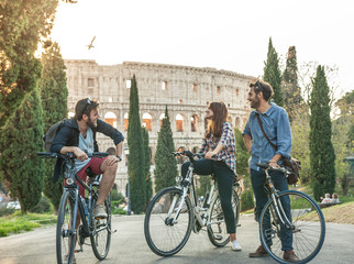 Wall Mural - Three young friends tourists with bikes in colle oppio park in front of colosseum on road with trees at sunset having fun talking laughing in Rome lens flare