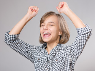 Close-up emotional portrait of caucasian little girl. Funny victory screaming teenager, on gray background. Lucky schoolgirl celebrating triumph, shouting with her hands up.