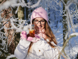 Wall Mural - Adorable happy young blonde woman in pink knitted hat scarf having fun drinking hot tea from mug snowy winter park forest in nature