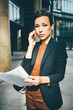 business women in black suit with paper documents on hands and speaking on the phone. Modern, contemporary glass building on background.