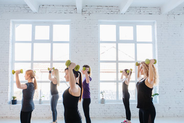 Wall Mural - Group of six sporty women workout with dumbbells in gym with white loft interior. Teamwork, good mood and healthy lifestyle concept.