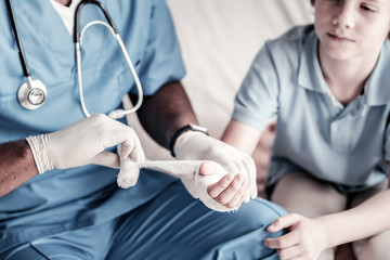 Wall Mural - Brave patient. Scaled up look on a millennial medical worker sitting next to a teenage patient and putting a bandage on his hand.