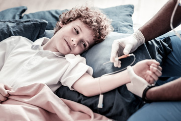 Wall Mural - I am not afraid. Smart teenage kid smiling slightly while looking at a male doctor putting a drip on his arm at hospital.