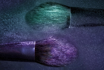 Two Brushes for makeup with purple and aqua make-up shadows in motion on a dark background.