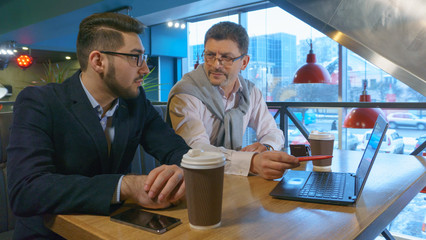 Two coworkers adult man in glasses and his young partner discusses a project with a laptop in a cafe