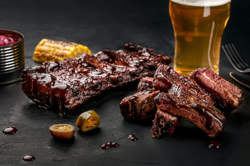 Pork ribs in barbecue sauce and a glass of beer on a black slate dish. A great snack to beer on a dark stone background. Top view with copy space