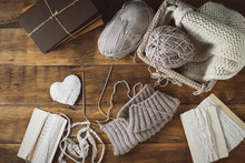 Light Gray Knits In A Basket And Knitting Needles, A White Knitted Scarf, A Heart. Set With White Ribbons On A Wooden Background. The Concept Of Handmade