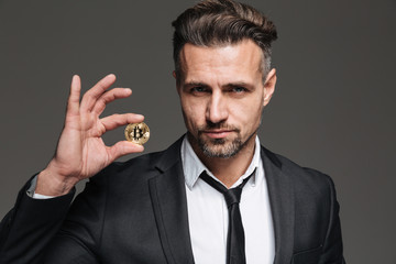 Canvas Print - Photo of successful mature man with brown hair in suit and tie demonstrating golden bitcoin on camera, isolated over dark gray background