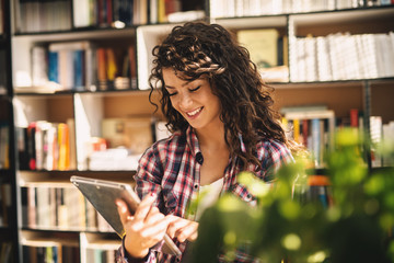 Attractive young satisfied student girl preparing lessons on the tablet while sitting in the library.