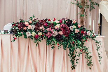 Rich bouquet of red, pink and white roses and greenery stands on the long dinner table for newlyweds table in a luxury restaurant