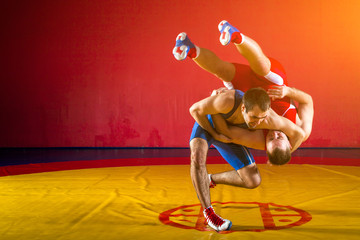 Two young men in blue and red wrestling tights are wrestlng and making a suplex wrestling on a yellow wrestling carpet in the gym