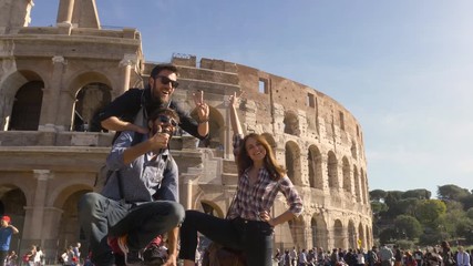 Wall Mural - Three young friends tourists standing on pedestal in front of colosseum in rome taking funny hilarious pictures posing with backpacks sunglasses happy beautiful girl long hair slow motion