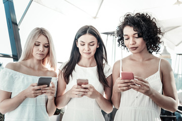 Wall Mural - Digital generation. Nice attractive young women standing together and using their smartphones while not paying attention to each other