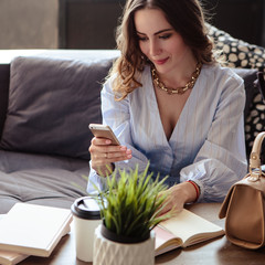 Wall Mural - Beautiful young woman with mobile phone and cup of coffee in stylish home interior. Freelancer business lady in her working place.