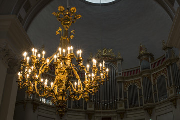 Detail of a luxury chandelier with golden decorations and hanging in a Christian cathedral. There are many lit electric candles that illuminate the church.