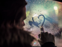 Side View Of Romantic Man Drawing Heart On Steamy Car Window Against Night Lights. 