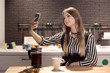 © Надежда Манахова - A young beautiful woman is sitting in the kitchen, drinking tea with a biscuit and taking a picture of herself, selfie. Modern dark interior. Generation Z.