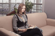 © Надежда Манахова - Beautiful young woman sitting on a beige couch with a book in her hands, waiting for her interior. The girl is dressed in a striped blouse and a black corset. Modern design. life style. Copy space.