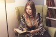 © Надежда Манахова - Beautiful young woman sits in a green armchair in the library and reads a book.