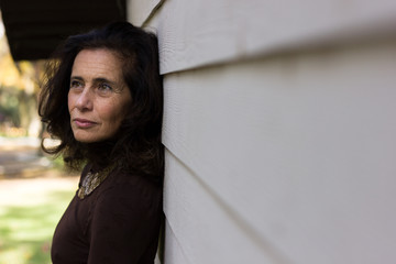 Serious mature woman on brown sweater and necklace leaning against white wooden plank wall. Concerned, worried look concept