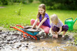 © MNStudio - Two funny little girls playing in a large wet mud puddle on sunny summer day. Children getting dirty while digging in muddy soil.