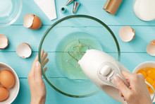 Female Hands Beating Egg Whites Cream With Mixer In The Bowl On Blue Wooden Table.
