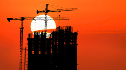 Silhouette of tower cranes in operation on top of the constructing building with background of the rising sun and golden sky